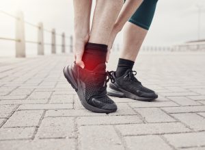 Jogger mit Knöchelverletzung auf gepflastertem Weg, hält schmerzenden Fuß in schwarzen Sportschuhen.